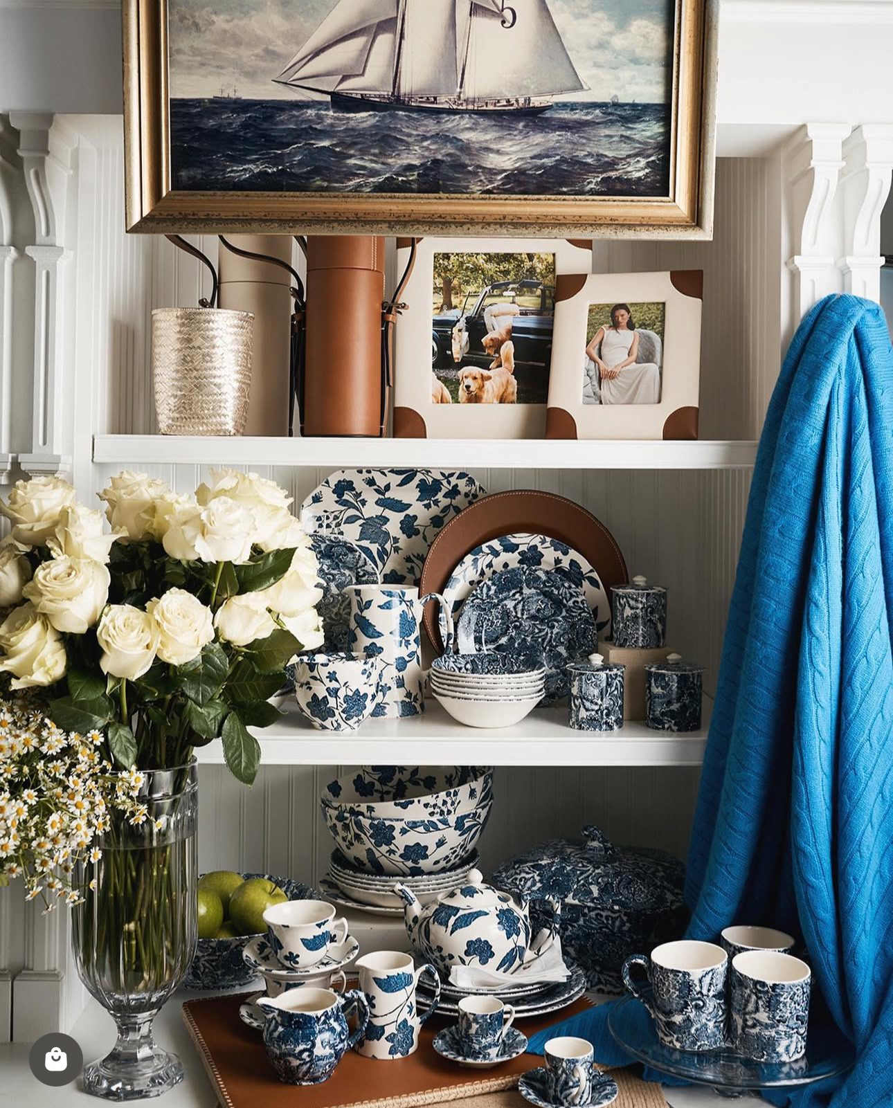 Elegant blue and white china displayed on shelves with fresh white roses and a blue curtain.